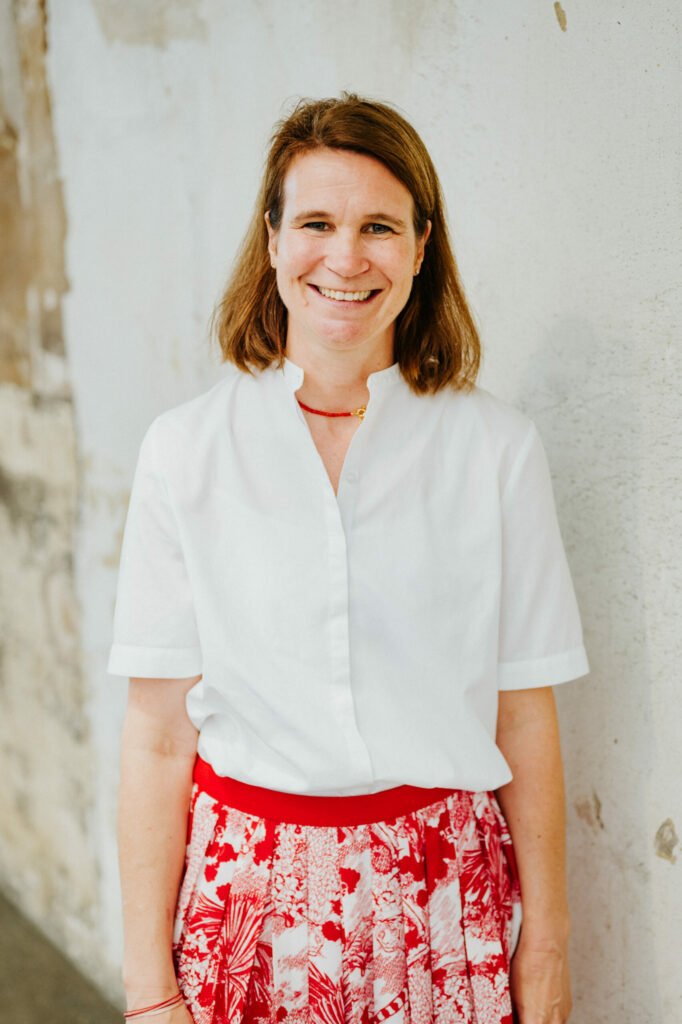Frau mit braunem Haar in weißer Bluse und rotem Rock vor heller Wand, lächelnd.