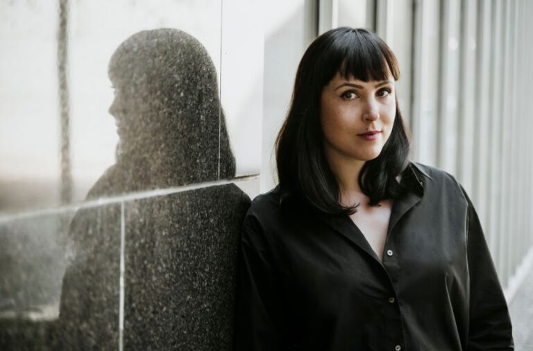 Frau mit dunklem Hemd und Haar steht neben einer Wand mit Spiegelung.