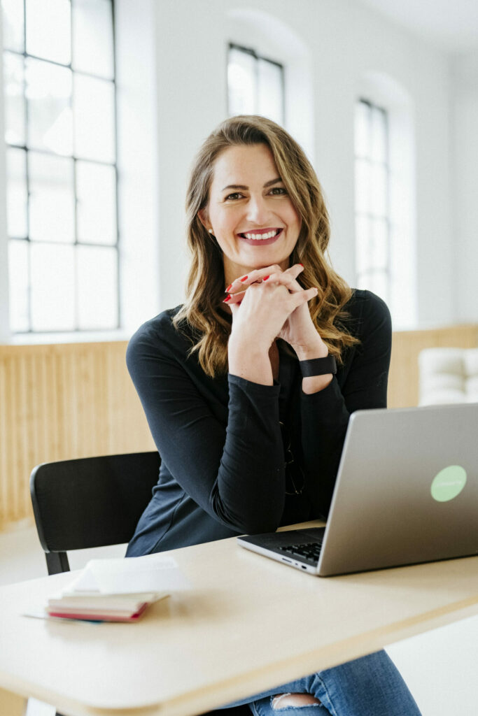 Frau mit Laptop am Tisch, schwarzes Oberteil, lange Haare, große Fenster im Hintergrund.