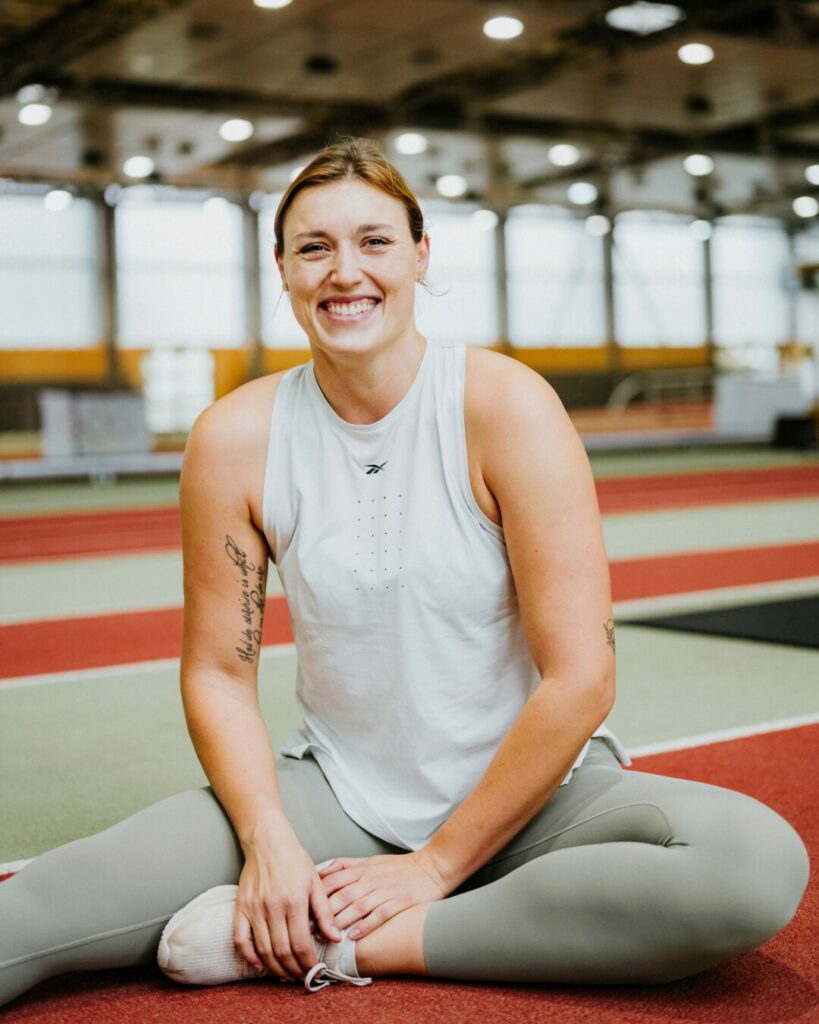 Lächelnde Person auf Sporthallenboden in weißem Top und grauen Leggings, Indoor-Sportbereich sichtbar.