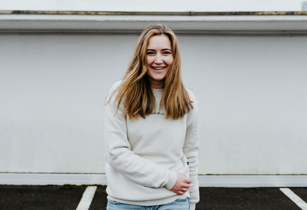 Person mit langen Haaren lächelt in hellem Pullover vor einer weißen Wand draußen.