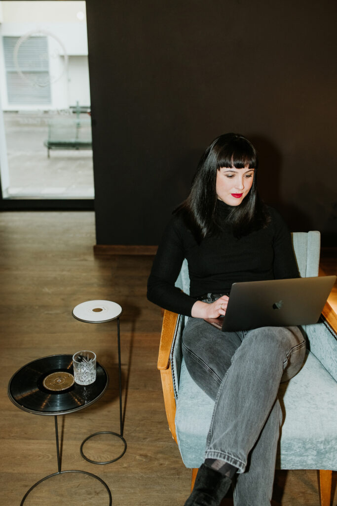 Person sitzt mit Laptop auf Sessel, Tisch mit Schallplatte und Glas, moderner Raum.