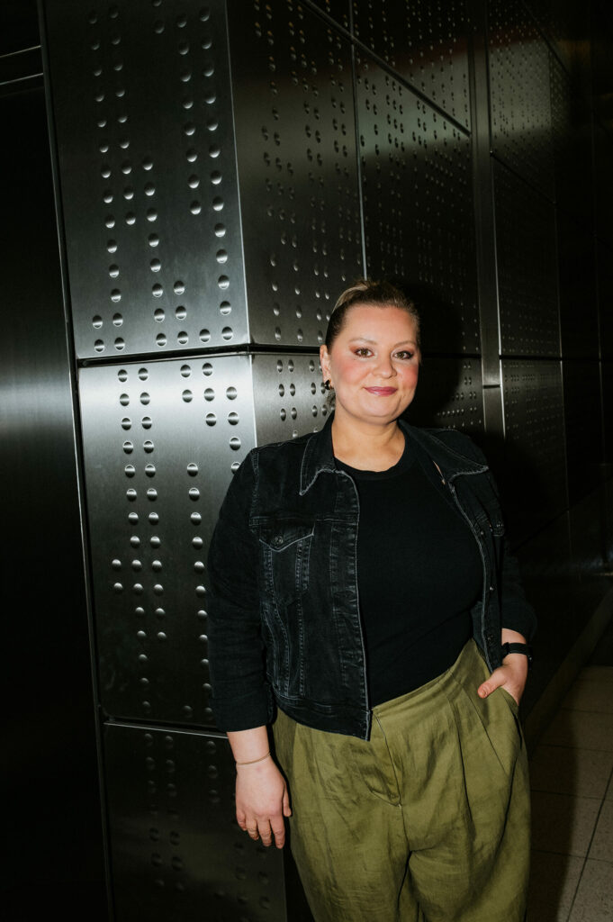 Person vor metallischer Wand, schwarze Jacke, olivgrüne Hose, regelmäßige Muster.