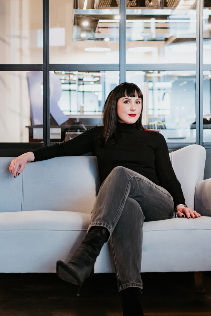 Person auf Sofa in modernem Raum mit Glaswänden, schwarzem Pulli, grauen Jeans.