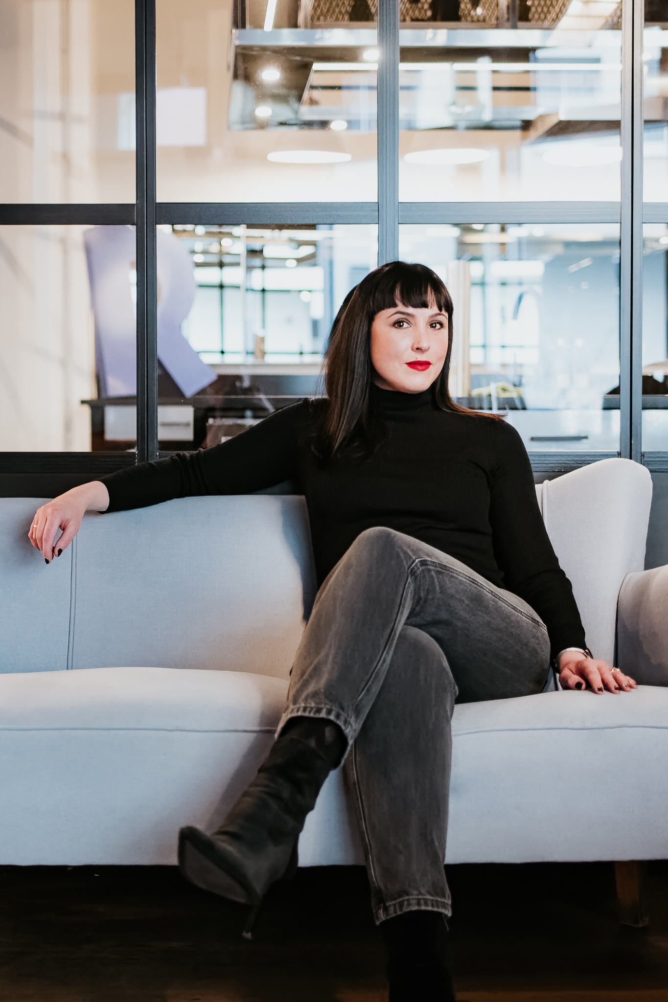 Person auf Sofa in modernem Raum mit Glaswänden, schwarzem Pulli, grauen Jeans.