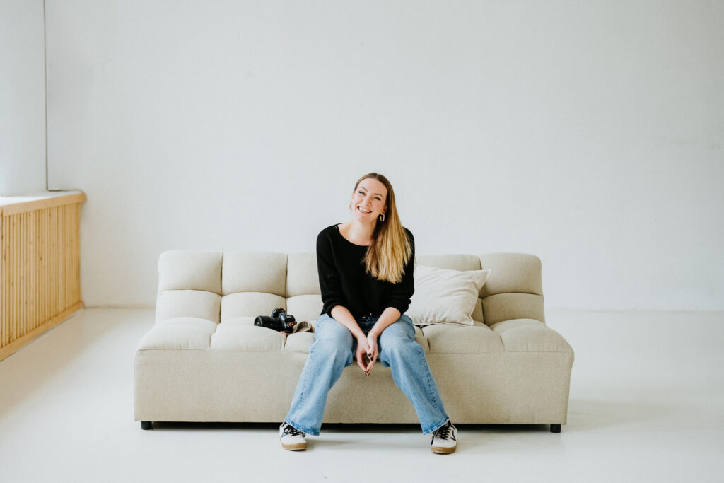 Person sitzt lächelnd auf cremefarbenem Sofa in hellem Raum mit Kamera, trägt Pullover und Jeans.