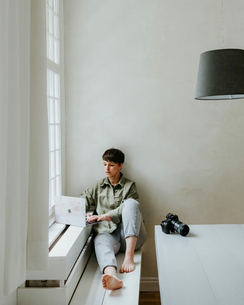 Person sitzt auf Bank am Fenster, arbeitet am Laptop, Kamera auf Tisch, heller minimalistischer Raum