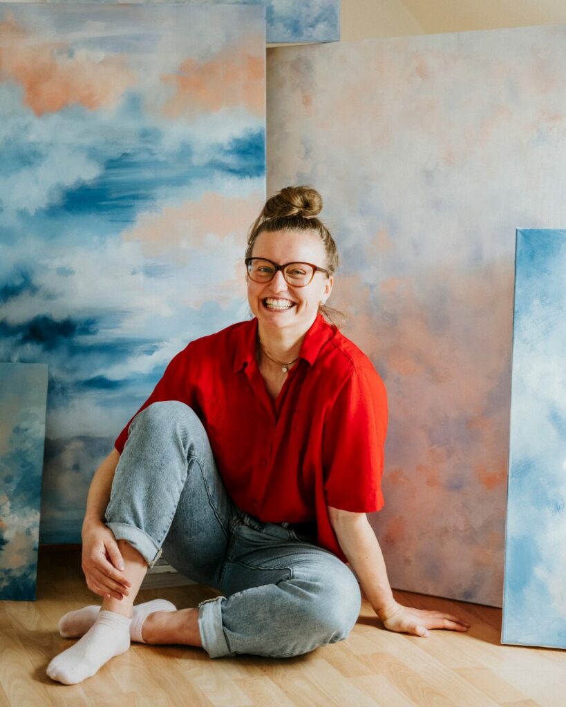 Lächelnde Person im roten Hemd, sitzt vor großen Kunstwerken mit blau-rosa Wolkenmustern.