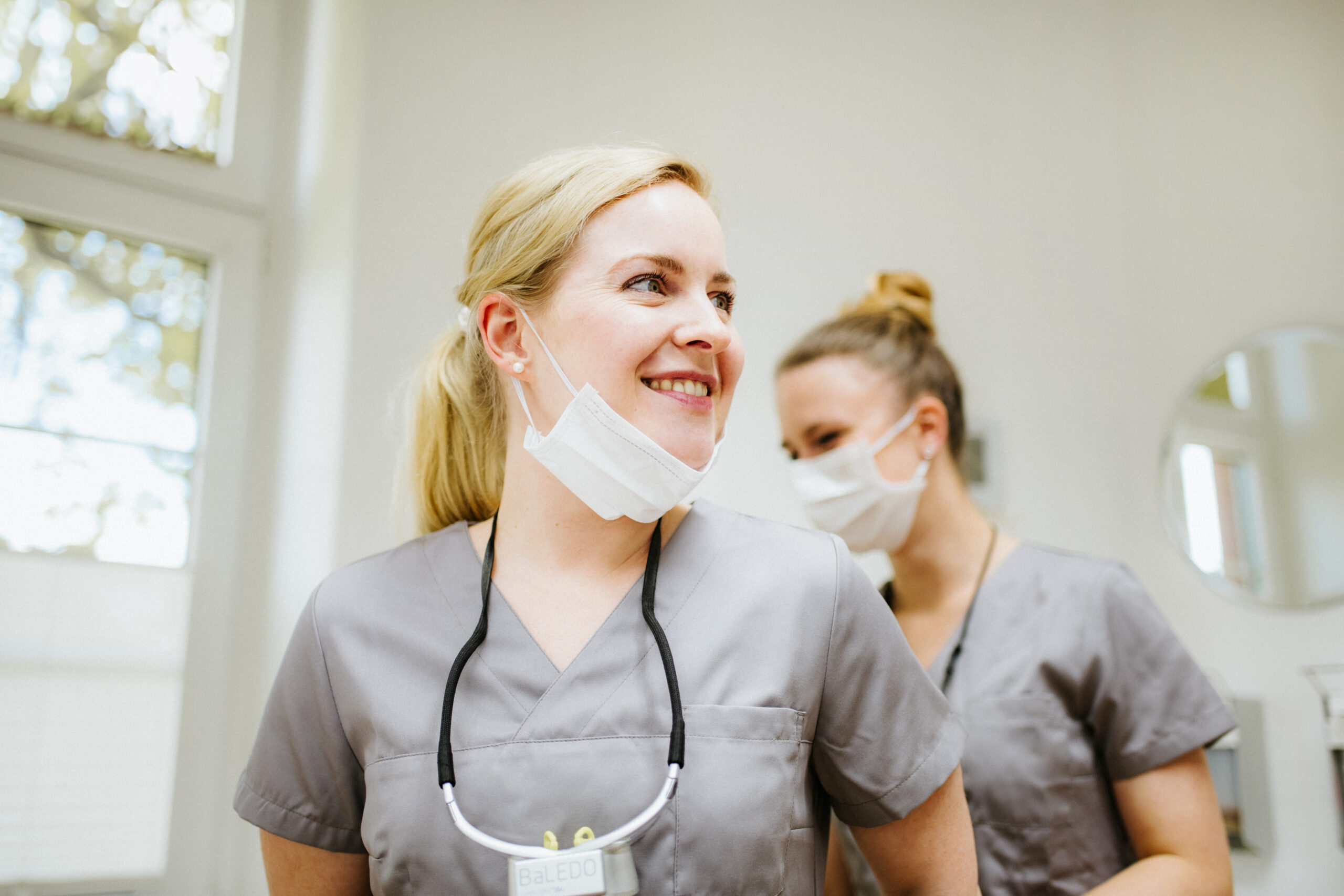 Zwei Frauen in grauen Kitteln mit Masken im hellen Raum, die sich unterhalten.