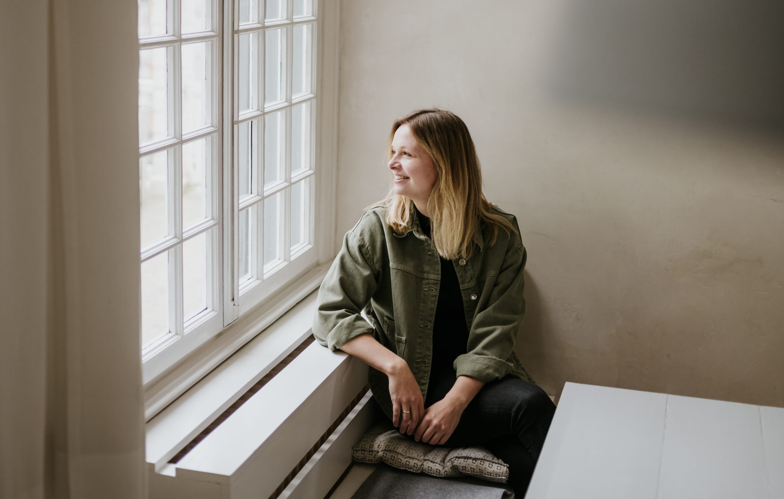 Person near window in green jacket, seated on cushion, smiling outward in bright, cozy setting.