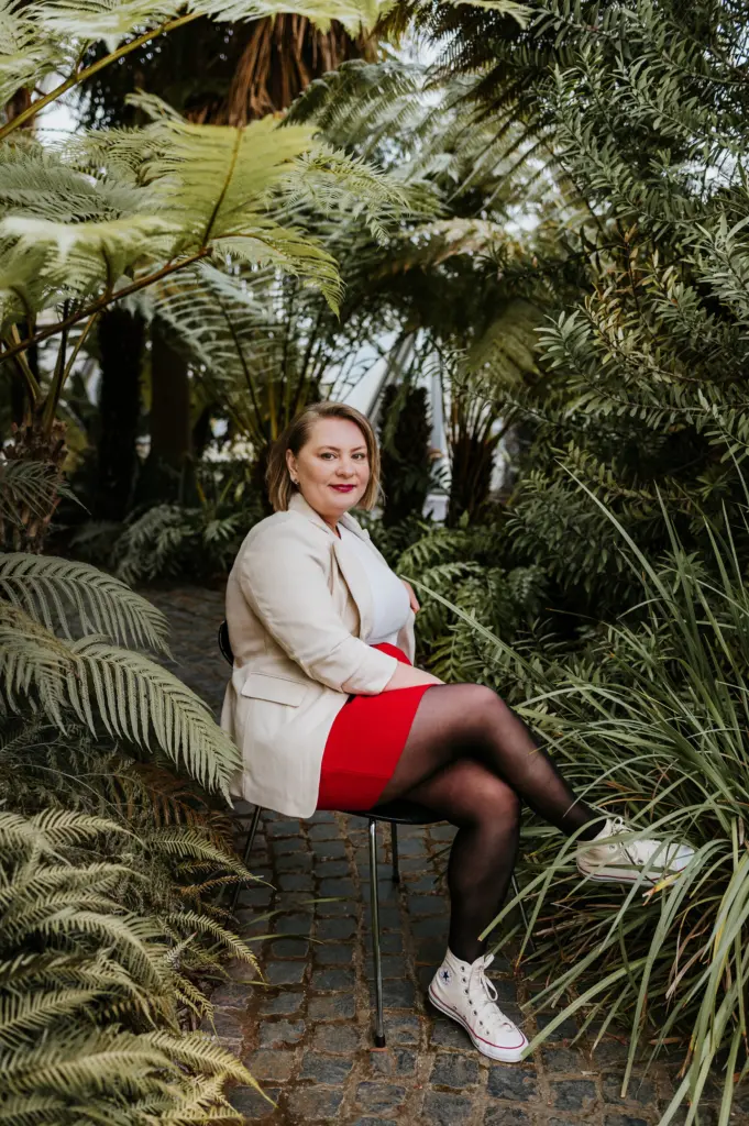 Frau in hellem Blazer, rotem Rock und weißen Sneakers sitzt auf Stuhl im grünen Gewächshaus.