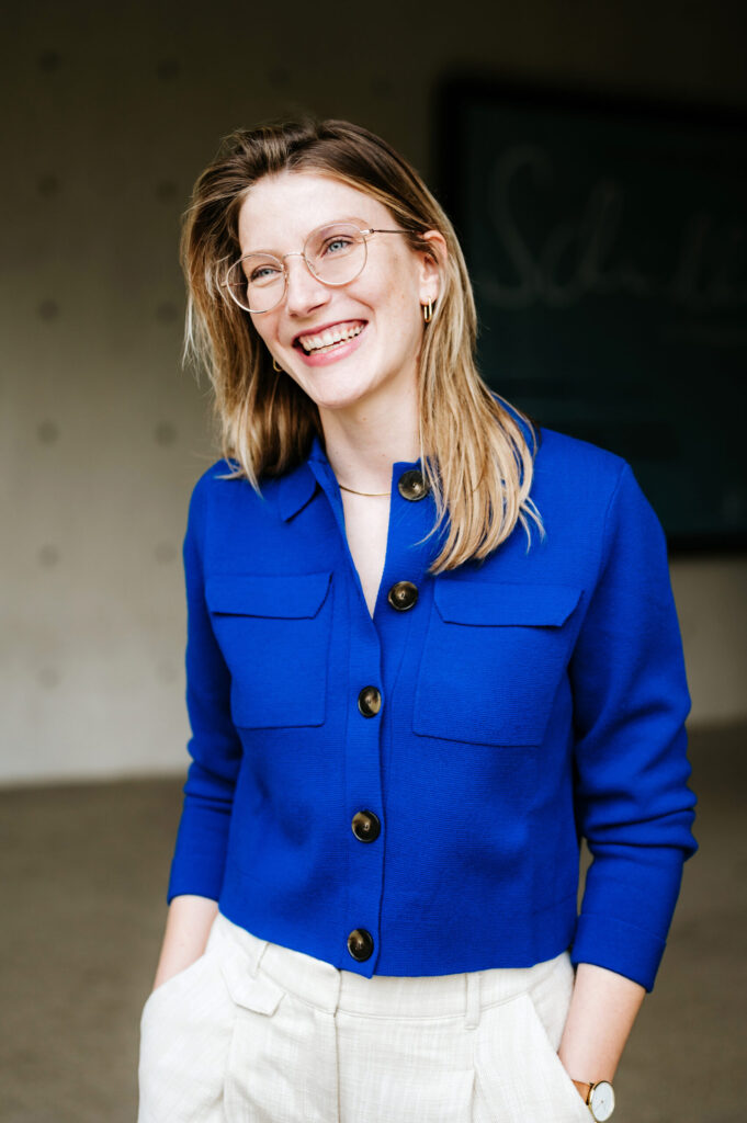 Frau mit blonder Schulterlänge, runder Brille, blauer Jacke und hellem Hose in Innenraum.