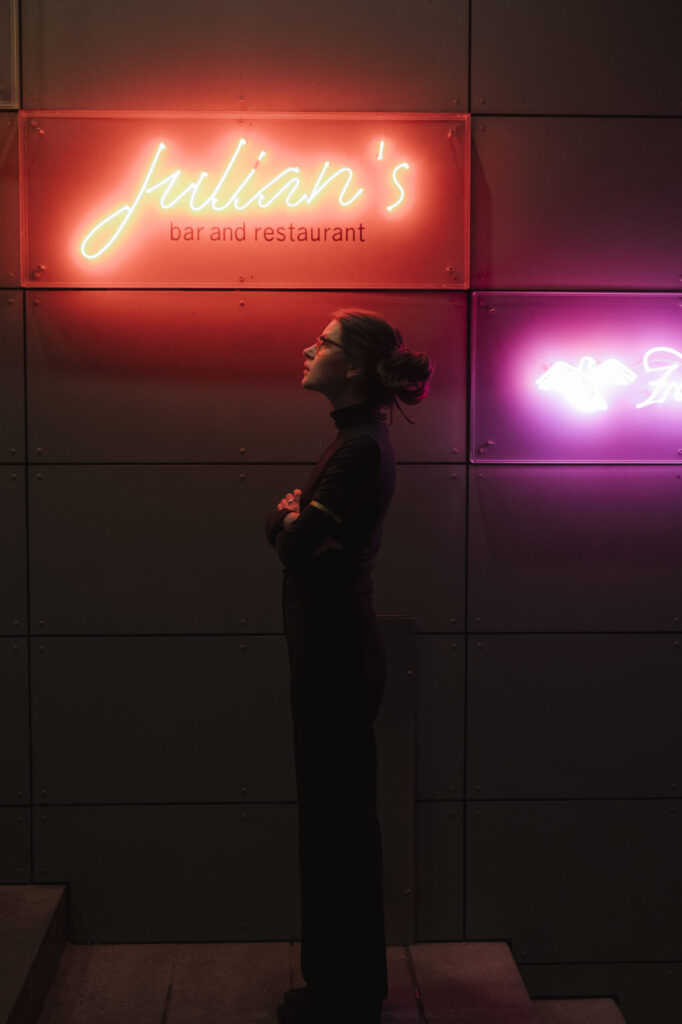 Frau in dunkler Kleidung steht vor Wand mit neonbeleuchtetem Schild Julian's bar and restaurant.