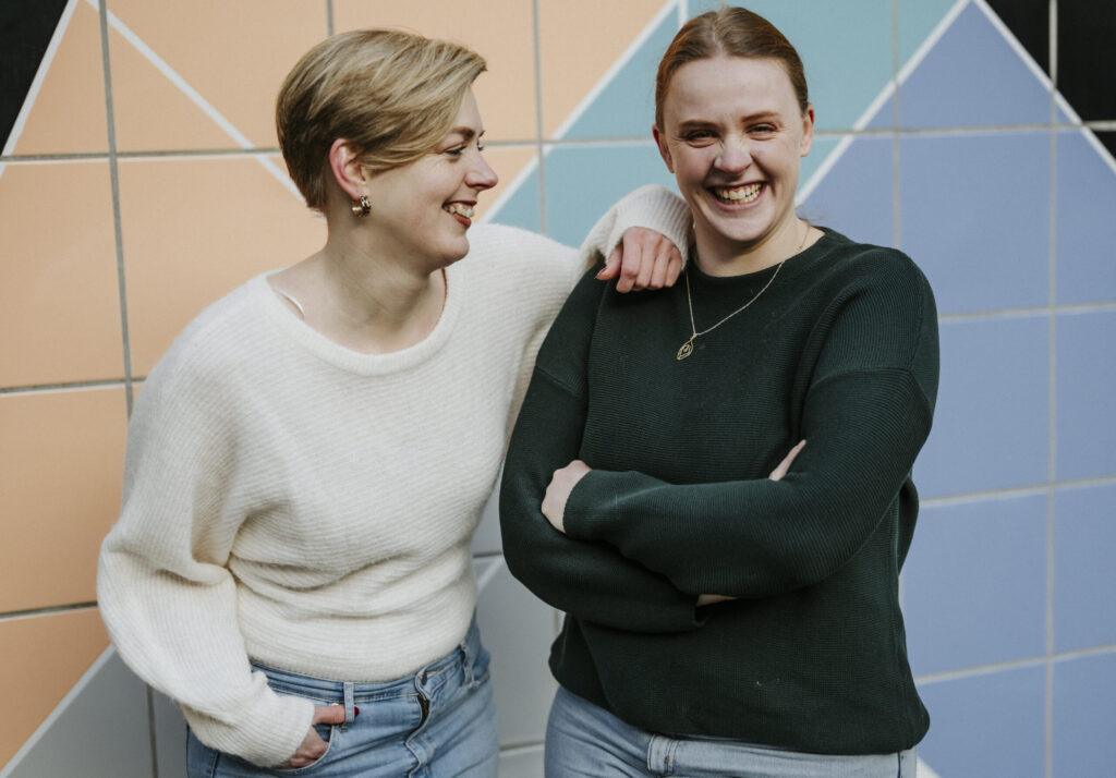 Zwei lachende Frauen in Jeans, eine mit weißem, eine mit grünem Pullover, vor bunter Wand.